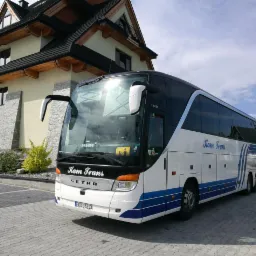 Autokar firmy Kam Trans zaparkowany przed budynkiem w stylu góralskim, widoczny znak informujący o przewozie dzieci.