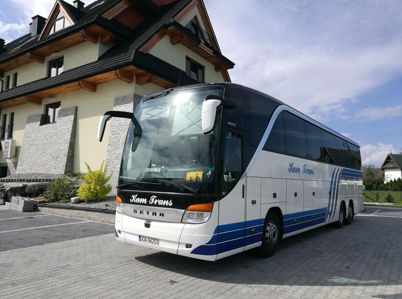 Autokar firmy Kam Trans zaparkowany przed budynkiem w stylu góralskim, widoczny znak informujący o przewozie dzieci.