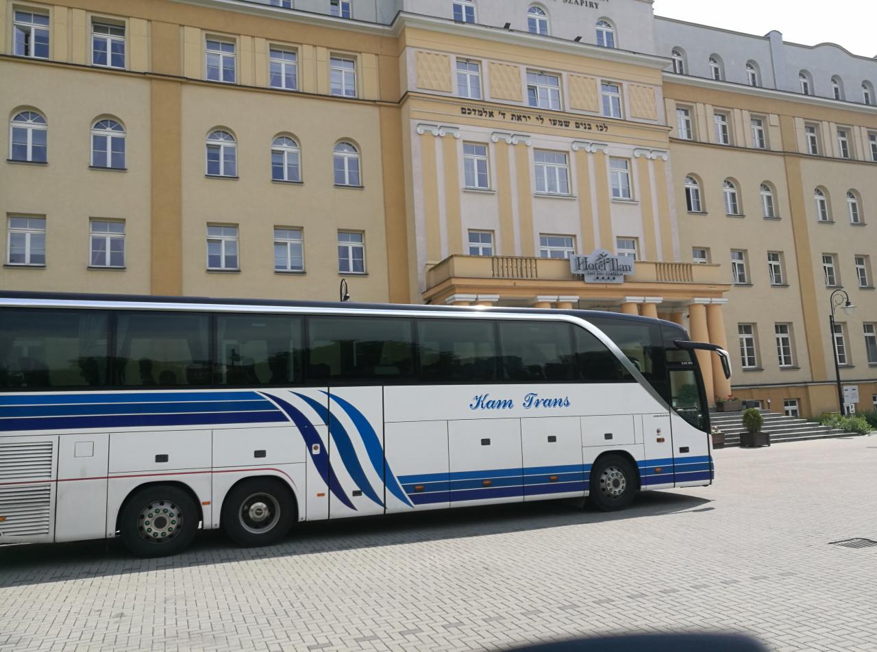 Autokar firmy Kam Trans zaparkowany przed hotelem z hebrajskim napisem na fasadzie.