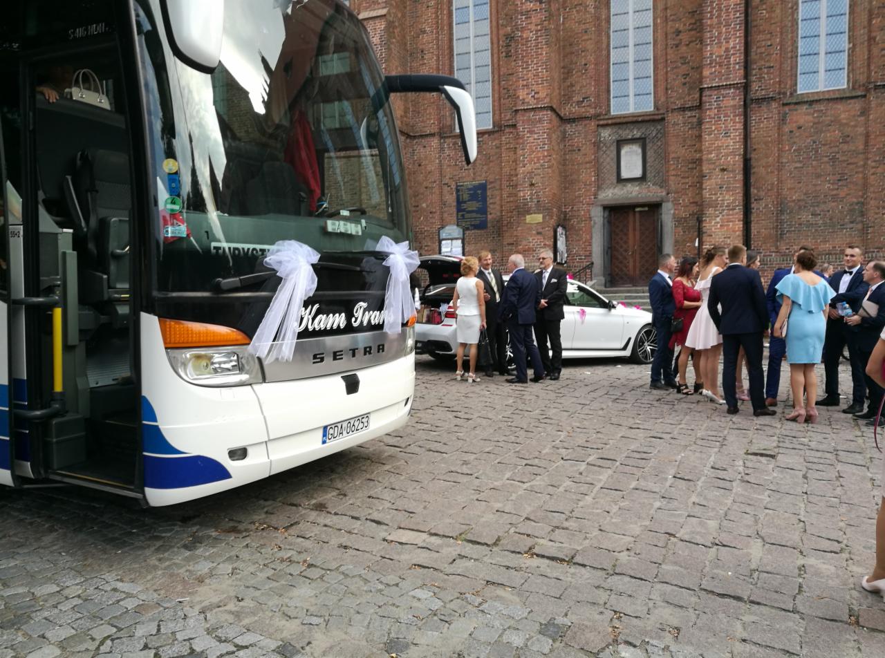 Dekorowany biały autokar z napisem 'Kam Trans' i rejestracją GDA-06253, ustawiony przed ceglanym budynkiem, obok białe auto z otwartym bagażnikiem i grupą ludzi w strojach weselnych.