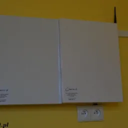 Dwa białe panele sterowania systemem alarmowym, zamontowane na żółtej ścianie nad podwójnym gniazdkiem elektrycznym; widoczna antena wystająca z prawego panelu i logo firmy na dole paneli.