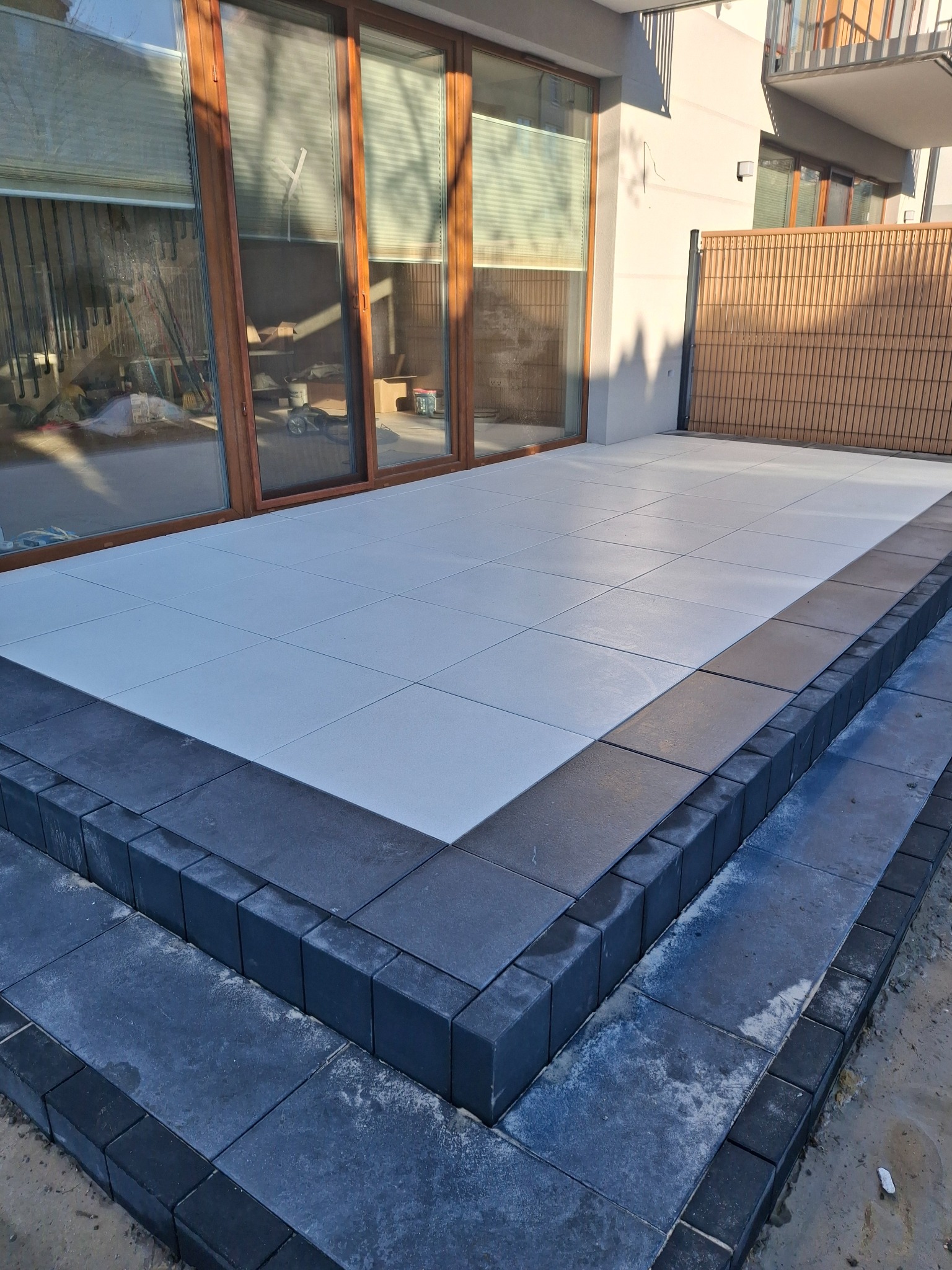 Nowoczesny taras z jasnych płyt, obramowany ciemną kostką brukową, prowadzący do dużych, przesuwnych drzwi tarasowych. Minimalistyczny design, jasne kolory, schludne wykończenie.