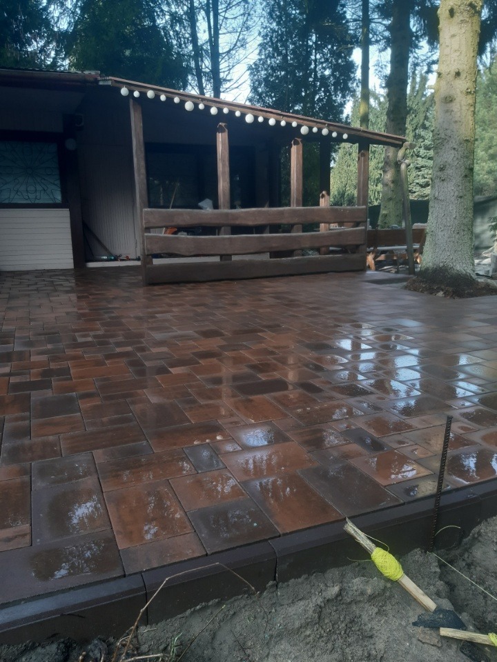 Ułożona, brązowa kostka brukowa na posesji z altaną i drzewem. Widoczne narzędzia brukarskie. Kostka lekko mokra, odbija światło. Krawężnik oddziela kostkę od ziemi.