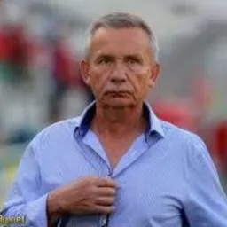 Portret starszego mężczyzny w niebieskiej koszuli w paski, z okularami w dłoni, na rozmytym tle stadionu sportowego, z widocznym logo w lewym dolnym rogu.