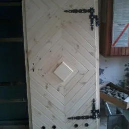 Drewniane drzwi wewnętrzne z dekoracyjnymi zawiasami i wzorem w jodełkę. Drzwi surowe, nielakierowane, z zaokrąglonym górnym brzegiem.