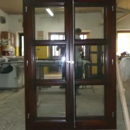 Ciemnobrązowe, drewniane, dwuskrzydłowe okno z szybami, ustawione w warsztacie. Widoczne refleksy i elementy wyposażenia warsztatowego w tle.