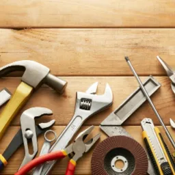 Różnorodne narzędzia, w tym młotek, poziomica, klucze, kombinerki i miarka, ułożone na drewnianym blacie, tworzące kompozycję roboczą.