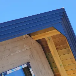 Niski kąt widzenia na narożnik budynku z widoczną konstrukcją drewnianą dachu i elementami betonowymi, pokryty ciemnoniebieską blachą trapezową.