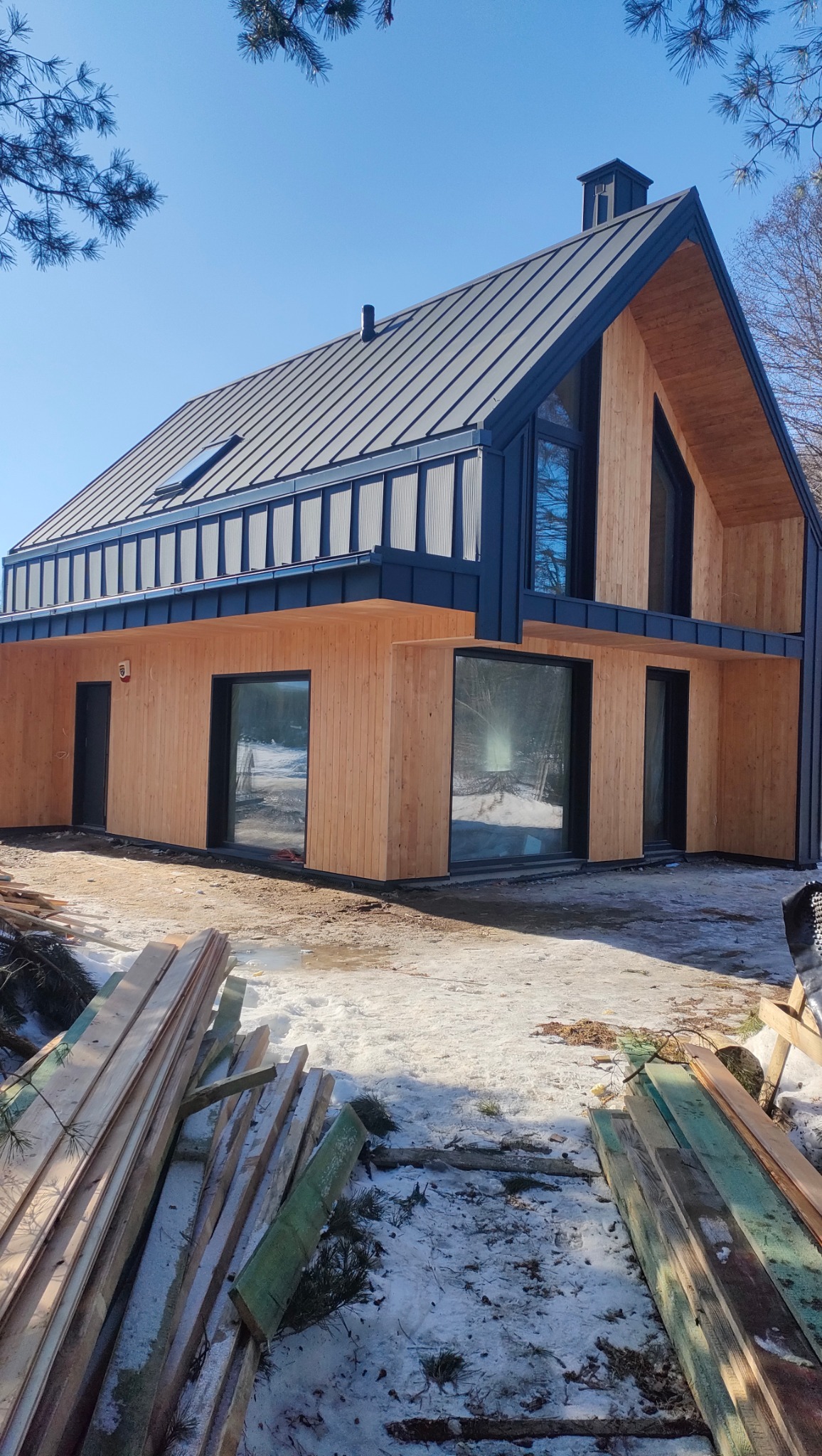 Nowoczesny dom z drewnianą elewacją i ciemnym, metalowym dachem. Widoczne deski na pierwszym planie sugerują trwające prace wykończeniowe.