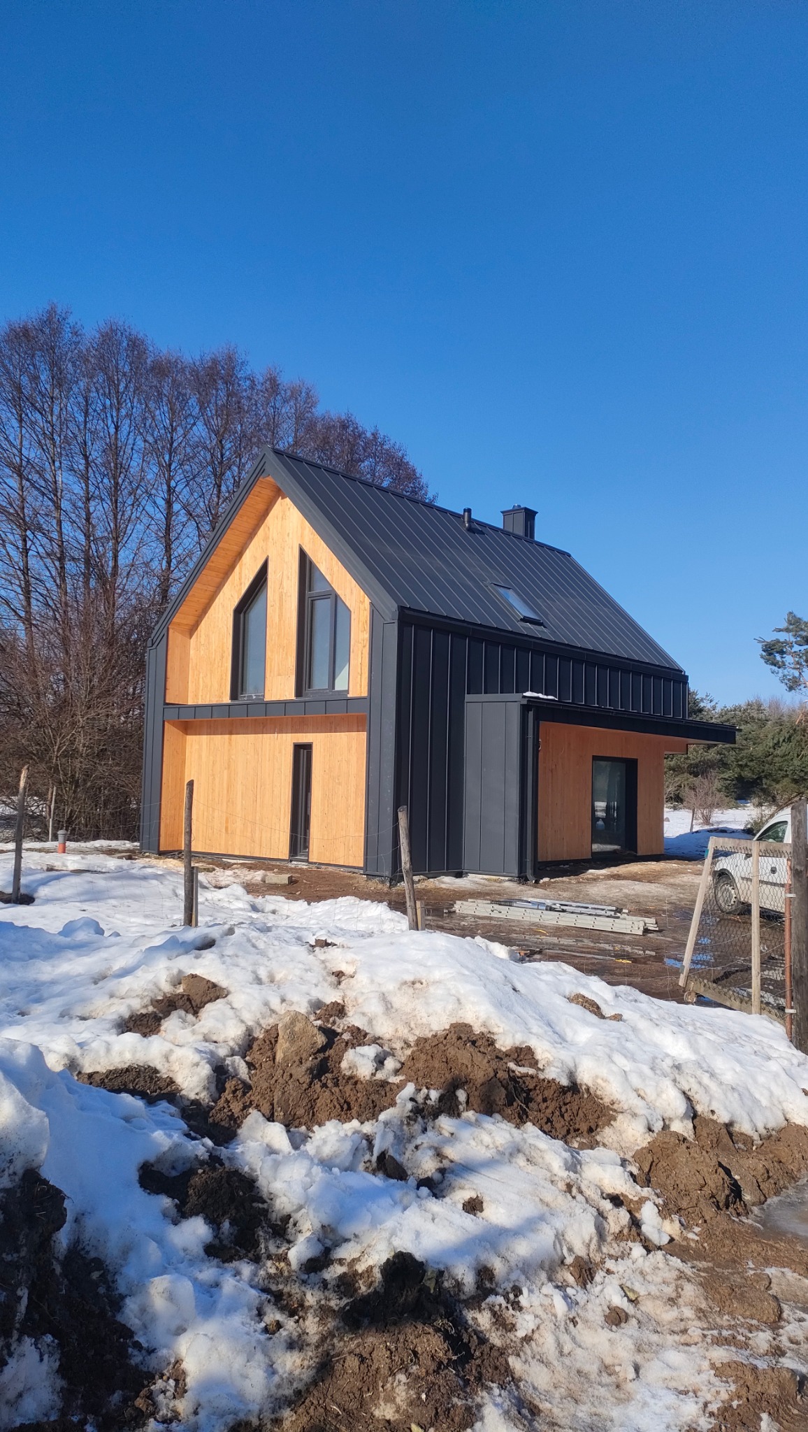 Nowoczesny dom z drewnianą elewacją i ciemnym dachem na tle błękitnego nieba i zimowego krajobrazu. Widoczne detale wykończenia i charakterystyczne okna.