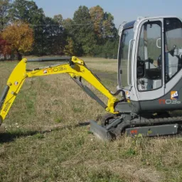 Żółto-szara mini koparka Wacker Neuson ET03 na gąsienicach, z kabiną i wysięgnikiem, stoi na trawiastym terenie. W tle drzewa i budynek.