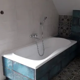 Biała wanna obudowana turkusowymi płytkami z efektem patyny, chromowana bateria z prysznicem, wzorzysta podłoga w stylu vintage, jasne płytki na ścianie w łazience na poddaszu.