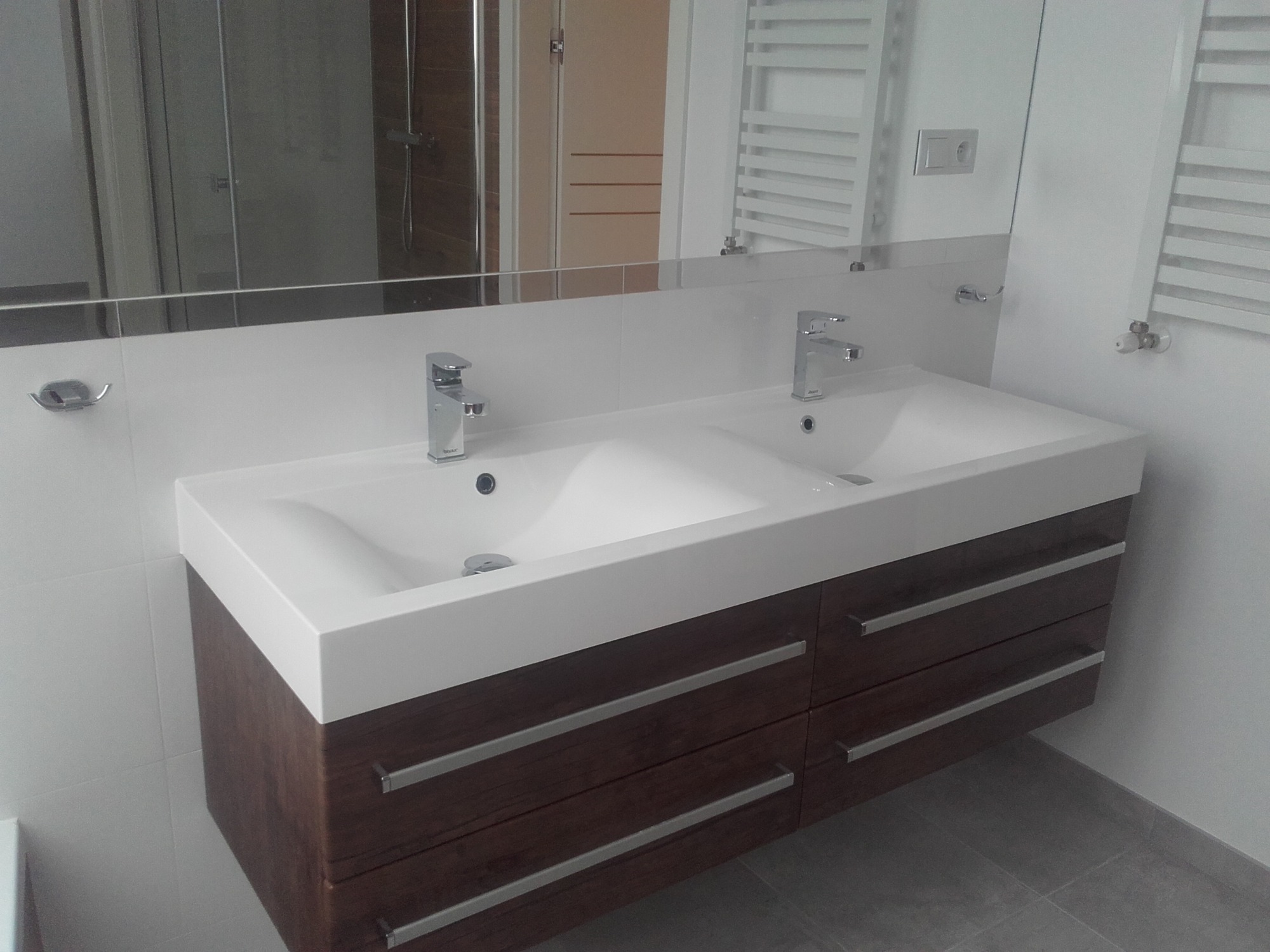 Nowoczesna łazienka z podwójną białą umywalką nablatową na szafce z ciemnego drewna i chromowanymi uchwytami, widok na lustro z odbiciem kabiny prysznicowej i grzejnika drabinkowego.