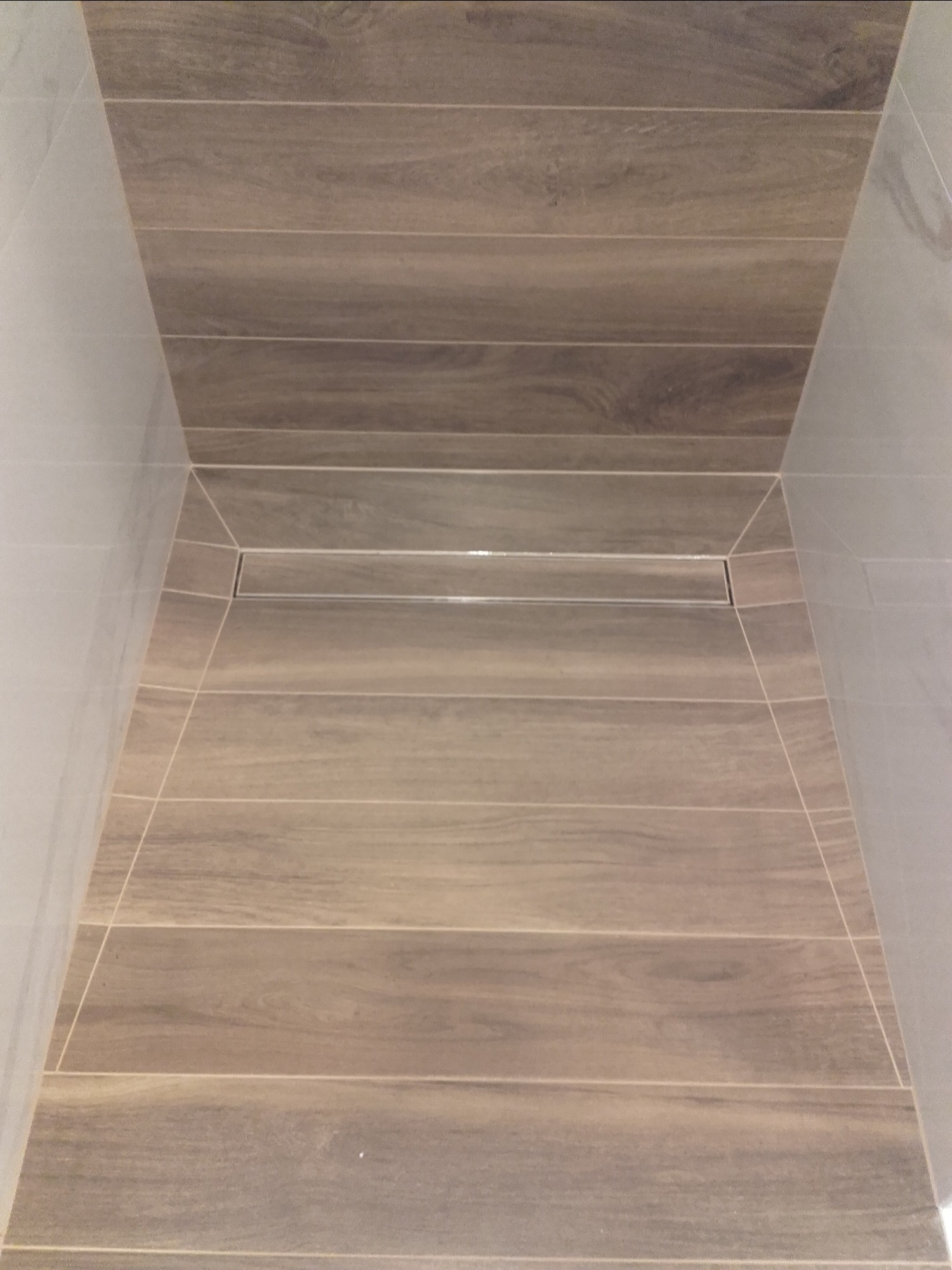 Wykończona kabina prysznicowa z odpływem liniowym, wyłożona podłużnymi płytkami imitującymi drewno, z jasnymi, gładkimi płytkami na ścianach.