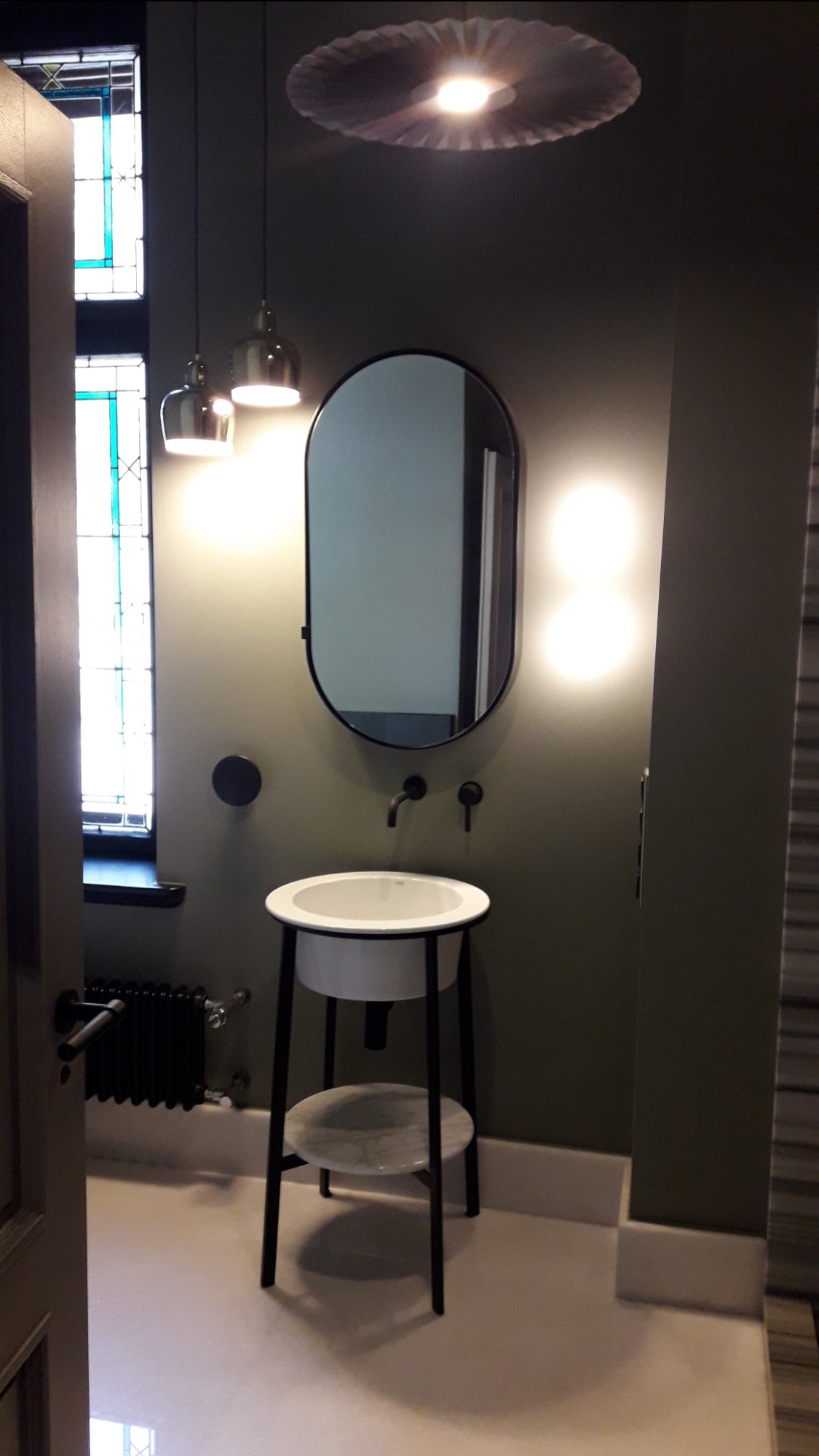 Nowoczesna łazienka z umywalką na metalowym stelażu, owalnym lustrem w czarnej ramie i designerskim oświetleniem, ściany w odcieniach zieleni.