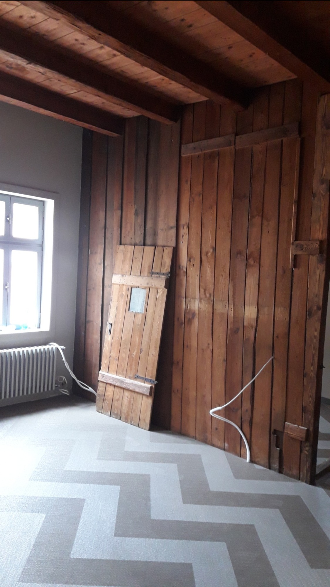 Wnętrze pokoju z drewnianą boazerią, starą drewnianą ścianą z drzwiami opartymi o nią, grzejnikiem pod oknem i podłogą w zygzaki.