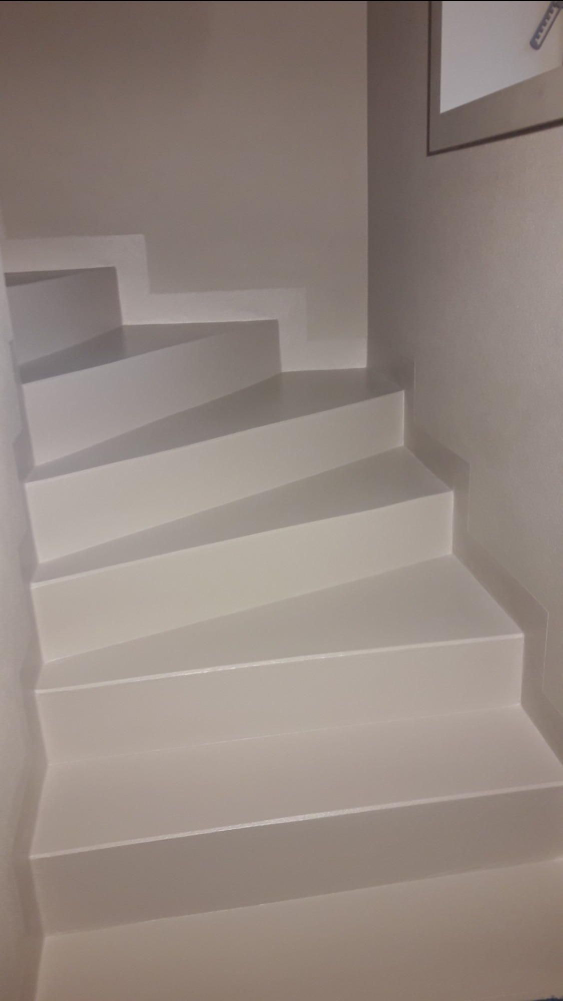 Nowoczesne, białe schody w wąskiej klatce schodowej, pomalowane farbą o delikatnym połysku, z widocznym fragmentem obrazu na ścianie.