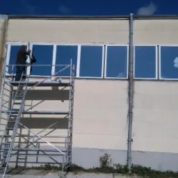 Pracownik na rusztowaniu montuje okno w budynku przemysłowym, widoczne odbicie chmur w szybach.
