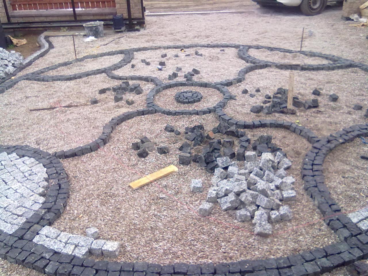 Układanie ozdobnego wzoru z kostki brukowej w trakcie realizacji, widoczne fragmenty ułożonego wzoru, stosy kostki brukowej oraz piasek.