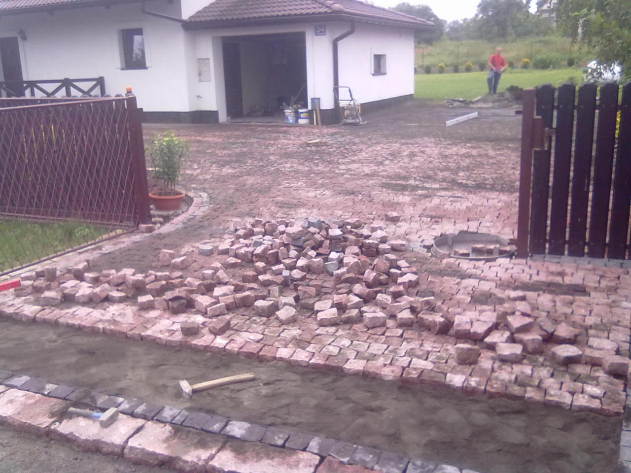 Niedokończona nawierzchnia z kostki granitowej przed domem, z hałdą kostek do ułożenia, widoczne narzędzia brukarskie i pracownik w oddali.