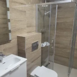 Nowoczesna łazienka z kabiną prysznicową, umywalką z szafką i podwieszaną toaletą, wykończona płytkami imitującymi drewno.