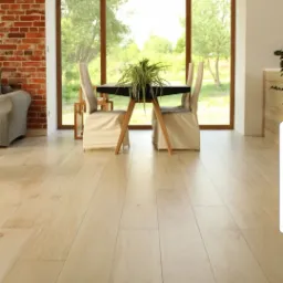 Jasne panele podłogowe w salonie z ceglaną ścianą, dużym oknem i stołem z krzesłami w pokrowcach.