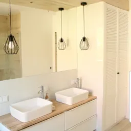 Nowoczesna łazienka z dwiema białymi umywalkami nablatowymi na drewnianym blacie, wiszącymi czarnymi lampami drucianymi i białą szafą z żaluzjowymi drzwiami.