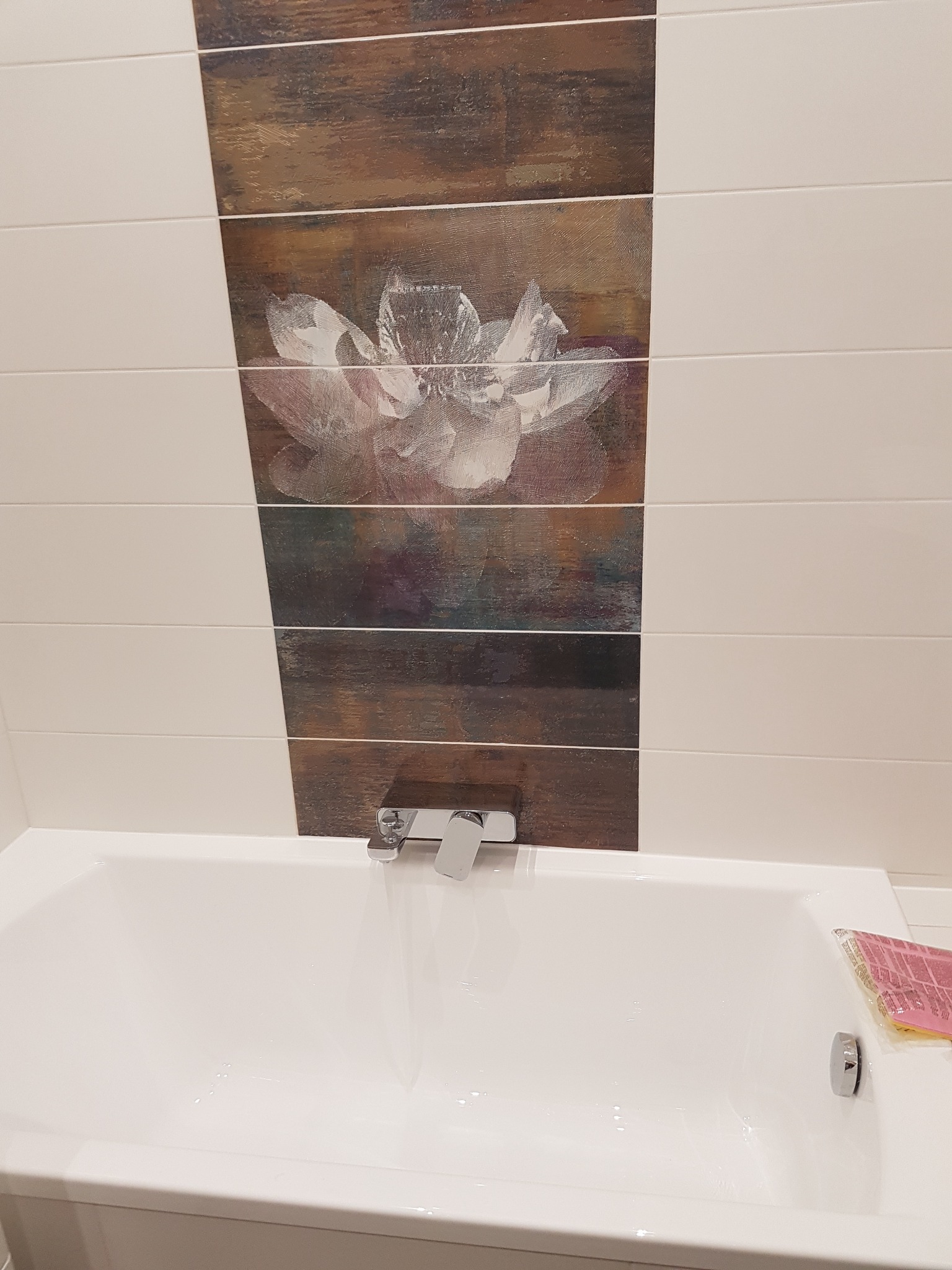 Biała wanna pod ścianą z poziomymi płytkami w kolorze białym i z motywem kwiatowym imitującym drewno.