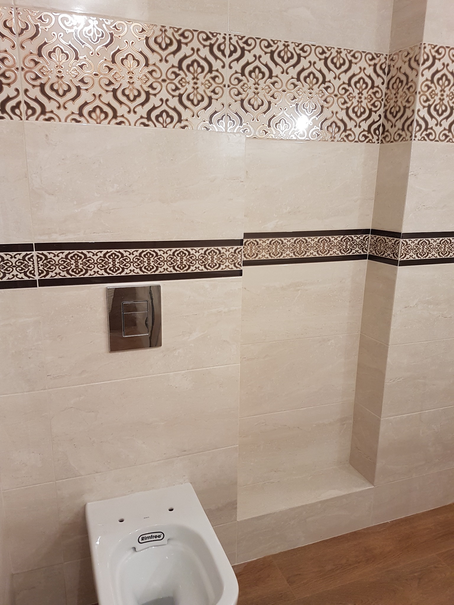 Beżowe płytki ścienne z dekoracyjnymi pasami wzorów nad zabudowaną spłuczką i częściowo widoczną miską WC Rimfree na tle drewnopodobnej podłogi.