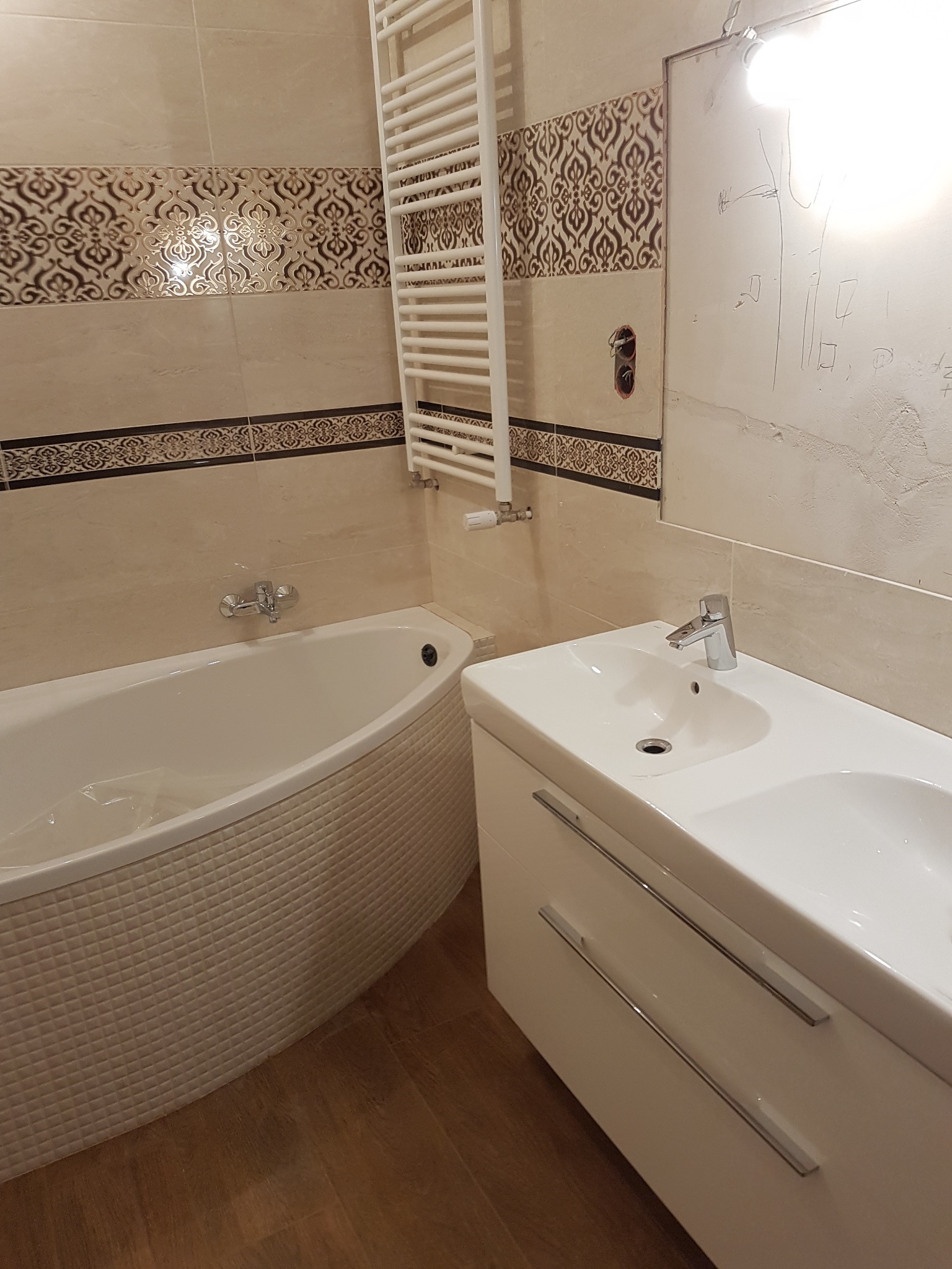 Wykończona łazienka z wanną obudowaną mozaiką, umywalką z szafką i grzejnikiem drabinkowym na beżowych płytkach z dekoracyjnym pasem.