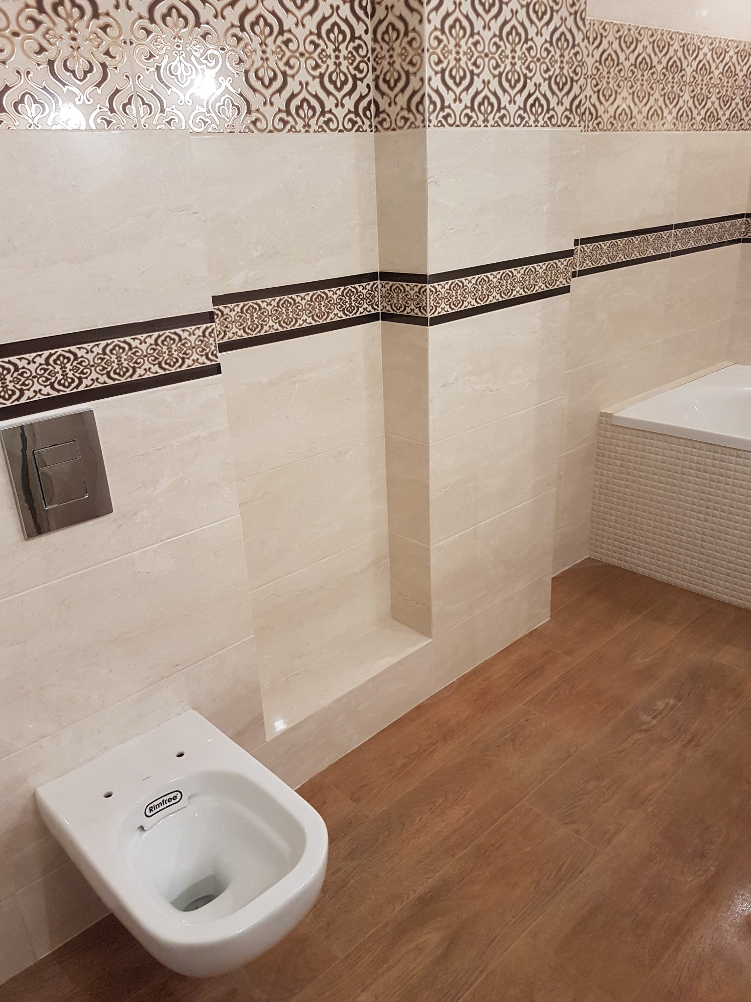 Nowoczesna łazienka z beżowymi płytkami zdobionymi ornamentami, wisząca miska WC marki Rimfree oraz drewnopodobna podłoga z paneli.