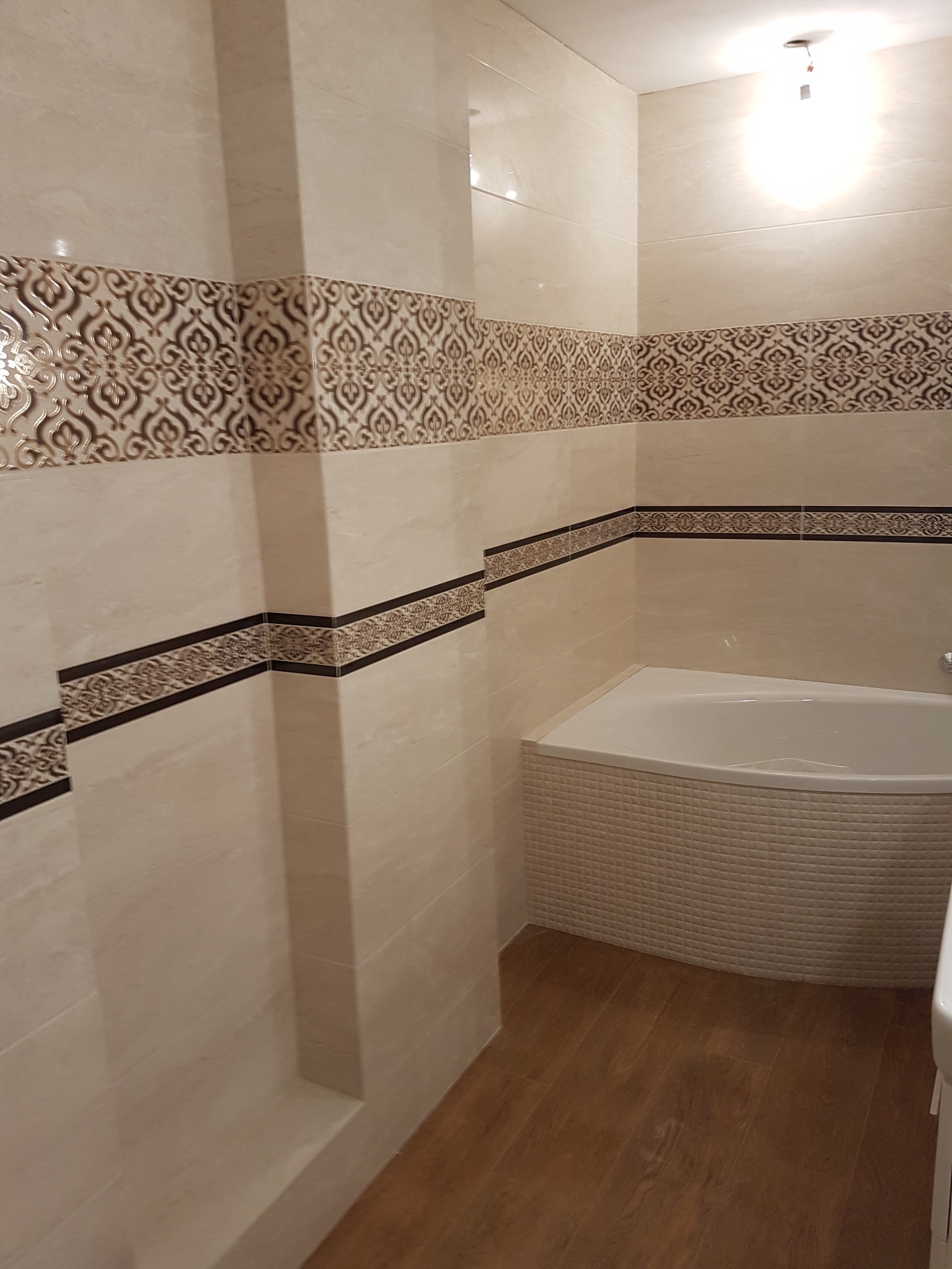 Wykończona łazienka z beżowymi płytkami i dekoracyjnymi pasami, drewnopodobna podłoga oraz narożna wanna obłożona mozaiką.