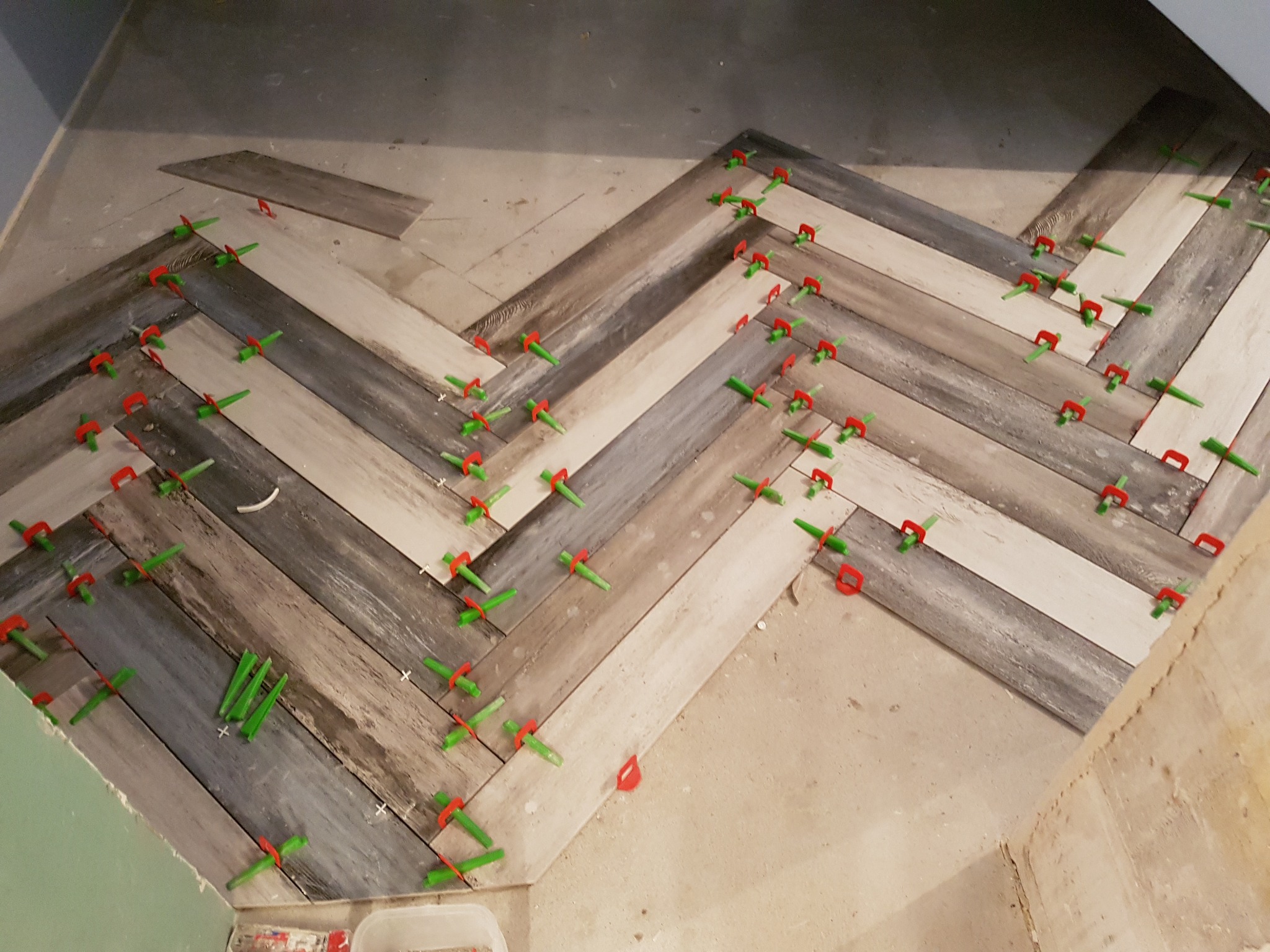 Układanie podłogi z drewnopodobnych paneli w jodełkę, z użyciem zielonych i czerwonych klinów dystansowych, widoczny surowy beton na podłodze i ściany w trakcie remontu.