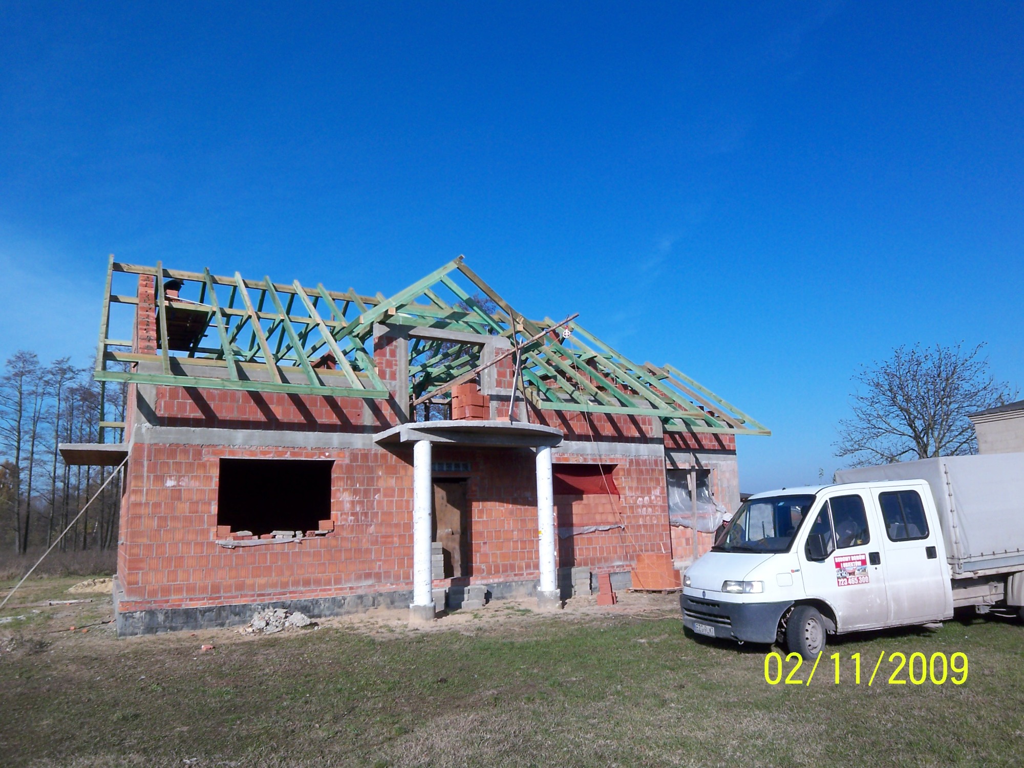 Niedokończony budynek z czerwonej cegły z drewnianą konstrukcją dachu, biały samochód dostawczy z reklamą firmy budowlanej zaparkowany obok, błękitne niebo w tle.