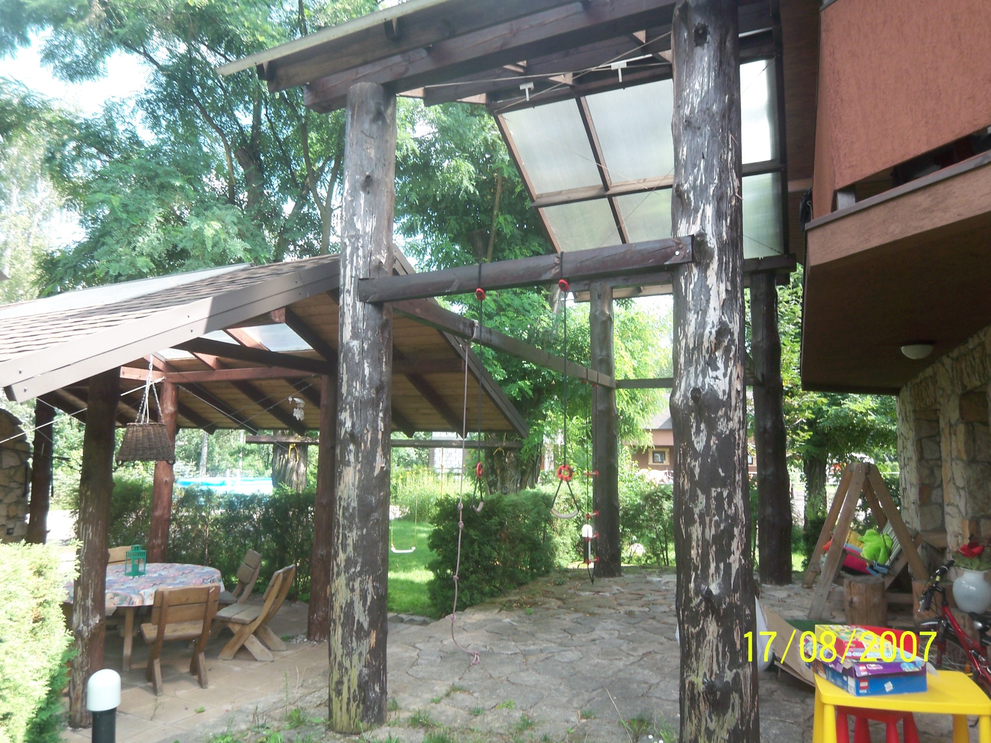 Zadaszona altana z drewnianych bali, stół z krzesłami pod zadaszeniem, huśtawki na sznurach, w tle widoczny basen i budynek mieszkalny.