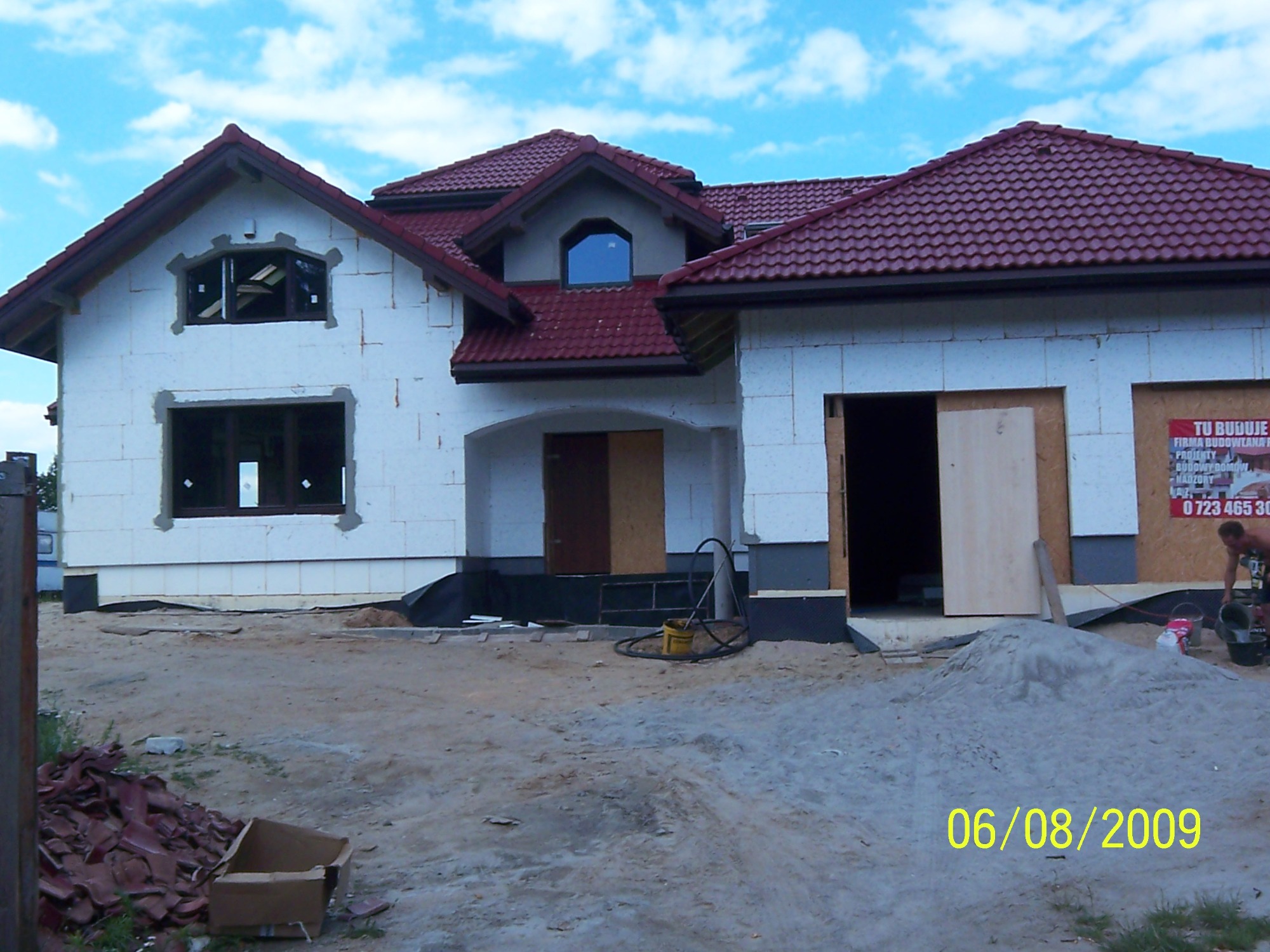 Surowy stan nowo budowanego domu z czerwonym dachem, widoczne bloczki z białego betonu komórkowego, niezabezpieczone okna, teren budowy z piaskiem i narzędziami, data na zdjęciu 06/08/2009.