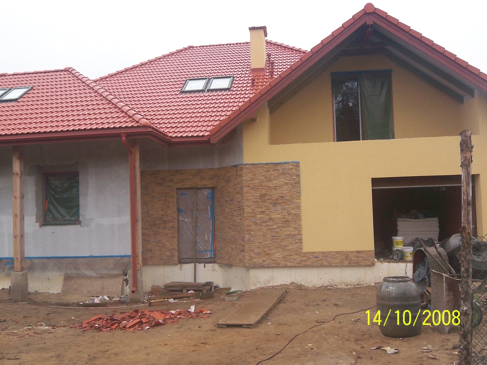 Niedokończona rozbudowa domu z nową elewacją z kamienia dekoracyjnego, widoczne okna zabezpieczone folią, gruz i materiały budowlane na placu budowy.