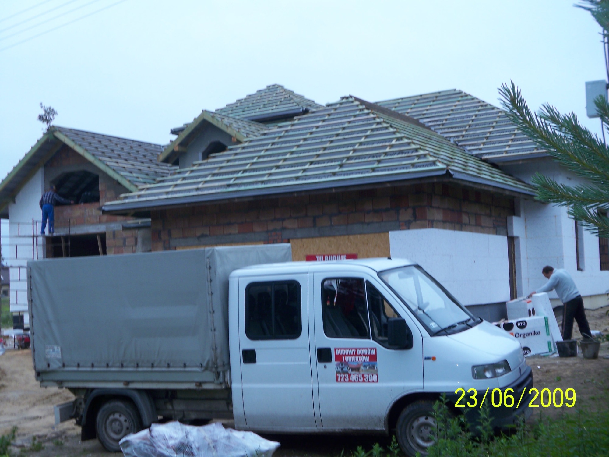 Budowa domu w trakcie realizacji: widoczny szkielet dachu z ułożoną dachówką, ściany z cegły i ocieplenie styropianem, oraz biała furgonetka z reklamą firmy budowlanej na tle pochmurnego nieba.