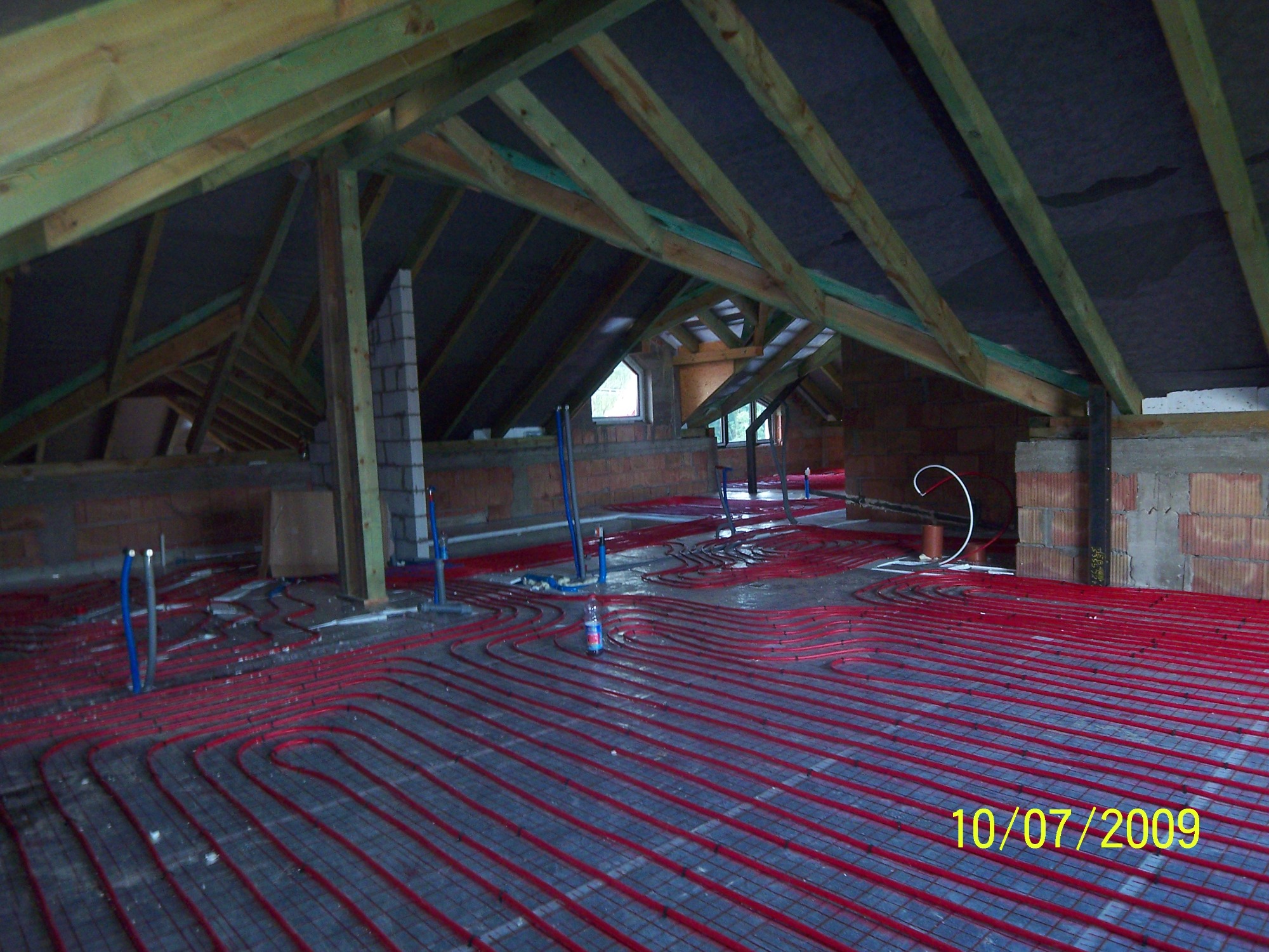 Montaż ogrzewania podłogowego z czerwonych rur na poddaszu w Zgierzu, widoczne elementy konstrukcji dachu i muru. Na podłodze leży butelka wody.