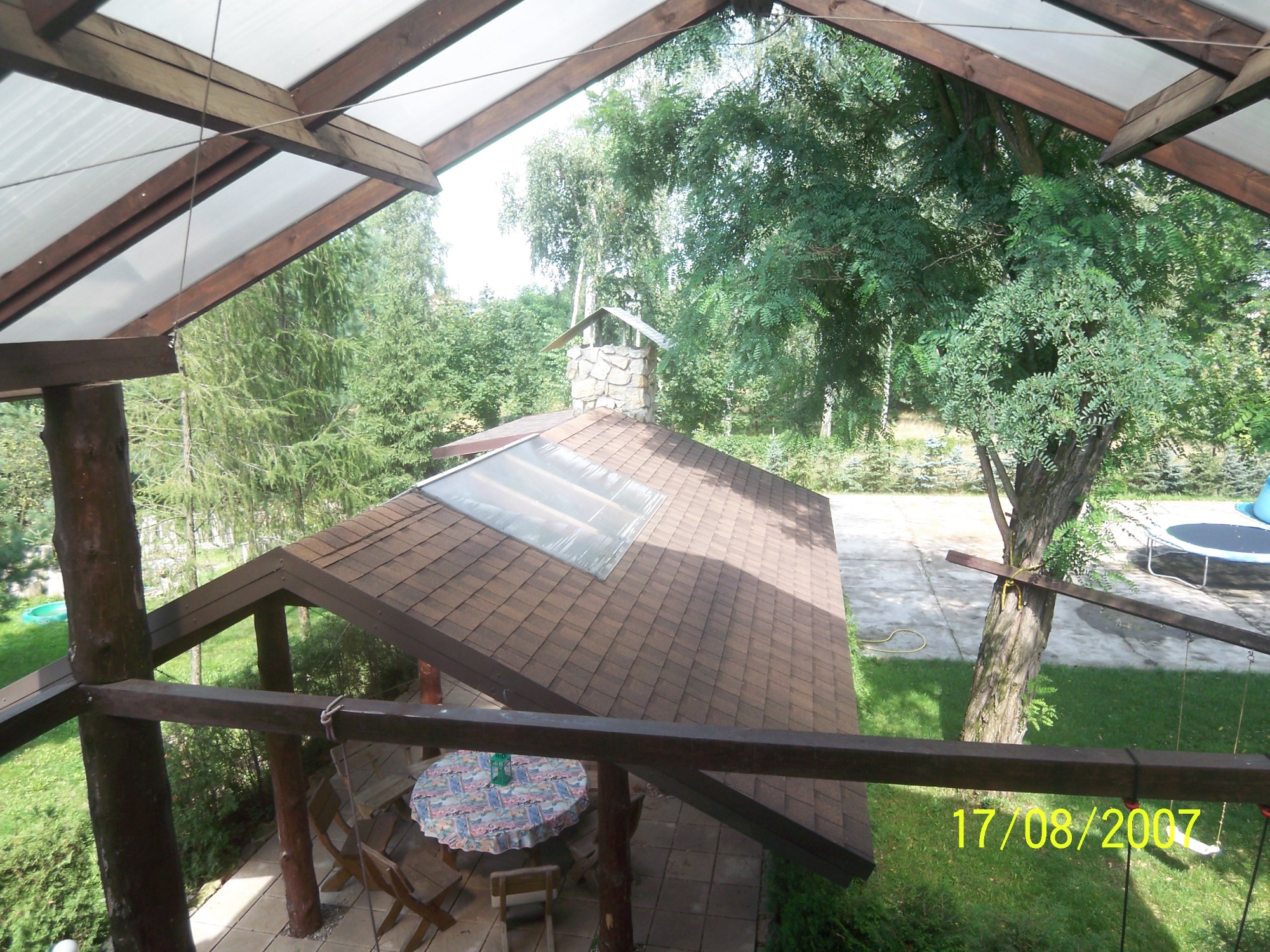 Widok z góry na drewnianą altanę z brązowym dachem krytym gontem bitumicznym i przezroczystym świetlikiem, otoczoną zielenią, z widocznym fragmentem placu zabaw i datą wykonania zdjęcia.