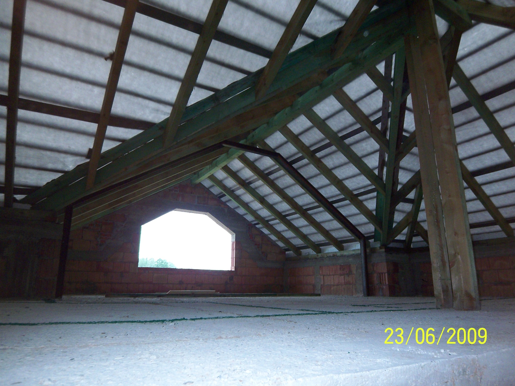 Konstrukcja dachu z drewnianych belek i membraną, widok z wnętrza poddasza, ściany z czerwonej cegły, okno z widokiem na zieleń, data 23/06/2009 w prawym dolnym rogu.