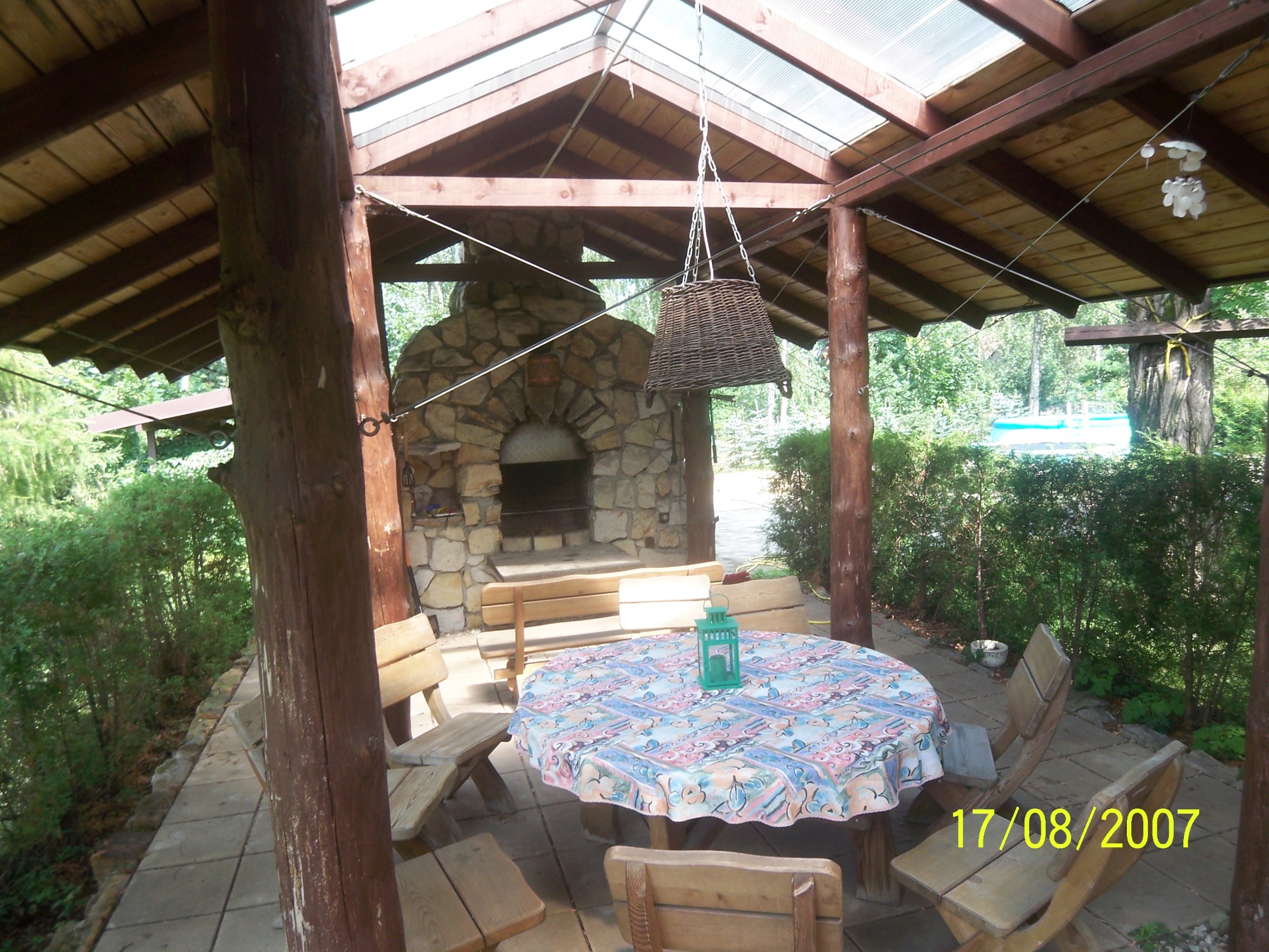 Zadaszona altana z kamiennym grillem, drewnianym stołem nakrytym wzorzystym obrusem i wiszącym koszem, otoczona zielenią.