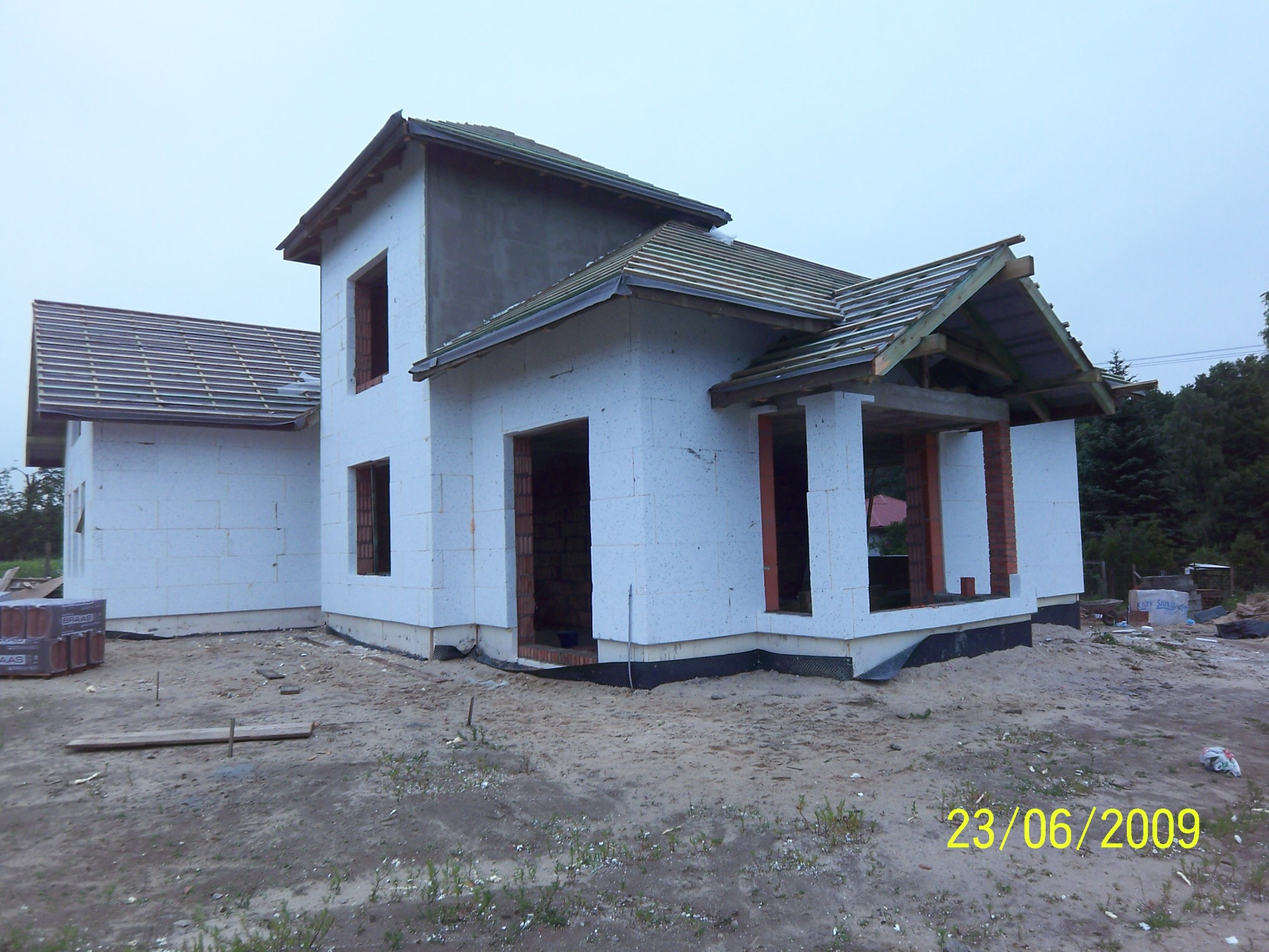 Surowy stan domu w trakcie budowy, ściany z białych bloczków, widoczne ceglane obramowania okien, na dachu ułożona dachówka, na gruncie piasek i resztki materiałów budowlanych.
