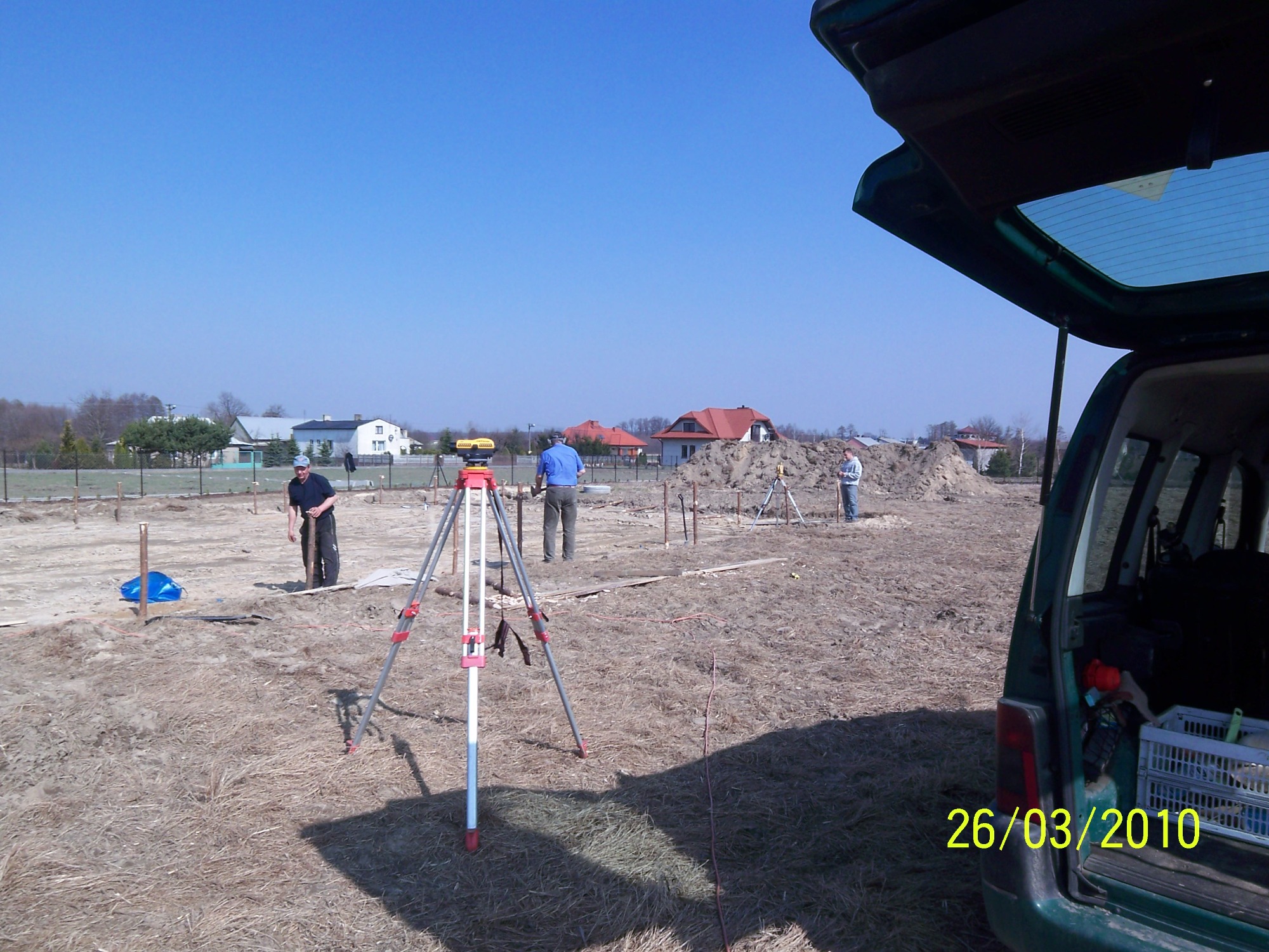 Geodeci wyznaczają punkty pod fundamenty domu na działce, widoczny otwarty bagażnik samochodu i sprzęt pomiarowy.