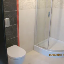 Wykończona łazienka z kabiną prysznicową narożną, toaletą wiszącą i płytkami w odcieniach bieli, szarości i czerwieni. Na ścianach widoczny dekoracyjny wzór. Ujęcie z perspektywy drzwi.