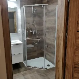 Nowoczesna kabina prysznicowa narożna z siedziskiem, brązowe płytki imitujące drewno na ścianach i podłodze, biała szafka umywalkowa w tle, widok zza otwartych drzwi.