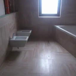 Jasna łazienka po remoncie z beżowymi płytkami, wiszącym bidetem i wanną pod oknem. Widoczny fragment instalacji. Minimalistyczny design.