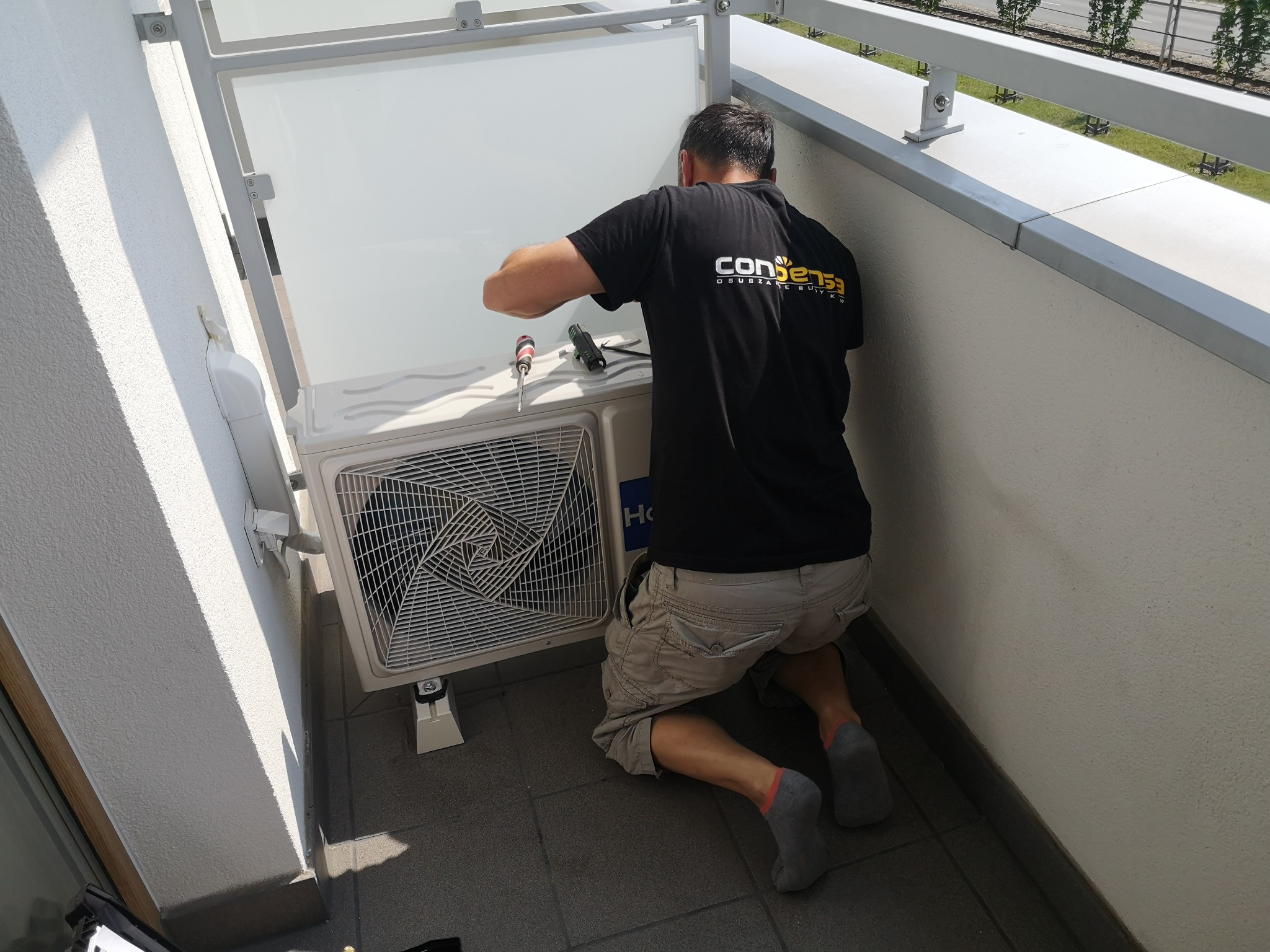 Instalator na balkonie, klęcząc, montuje zewnętrzną jednostkę klimatyzacji Haier, używając śrubokręta. Widoczne narzędzia i logo firmy na koszulce.