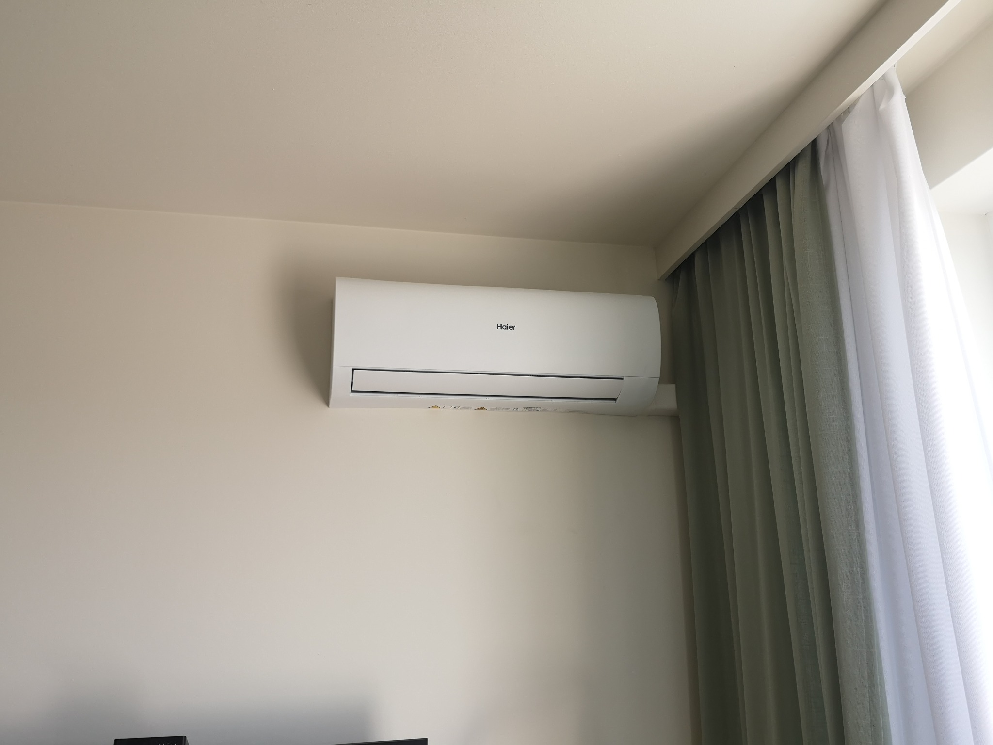 Biała jednostka klimatyzacji Haier zamontowana na jasnej ścianie w pomieszczeniu z zasłoniętym oknem.