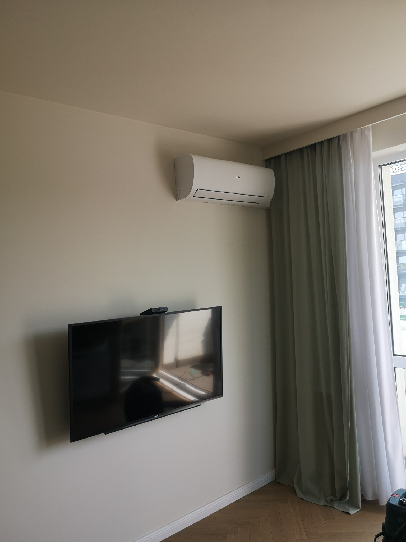 Biały klimatyzator Haier zamontowany nad telewizorem Sony w jasnym pokoju z zasłonami i widokiem na budynek za oknem.